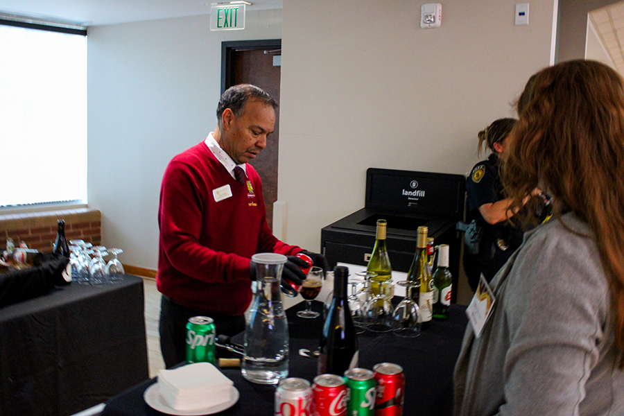 HILP Dinner 2025-2026 / A man in a red sweater serves drinks at a bar setup, with bottles of wine, soda cans, and glassware. A woman waits nearby. Bright, casual setting.