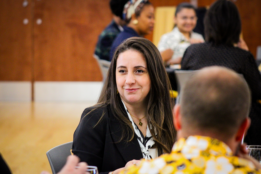 HILP Dinner 2025-2026 / A woman with long hair and a black jacket smiles while seated at a table in a lively conference room. Other attendees are visible in the blurred background.
