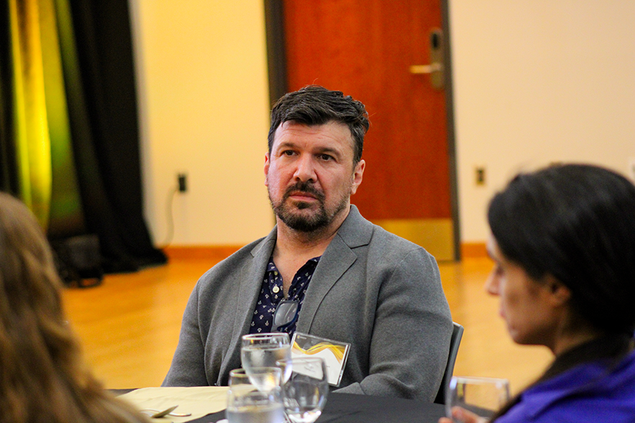 HILP Dinner 2025-2026 / Man seated at a formal event, wearing a gray blazer and patterned shirt, looking contemplative. Others are present, indoor setting with warm lighting.