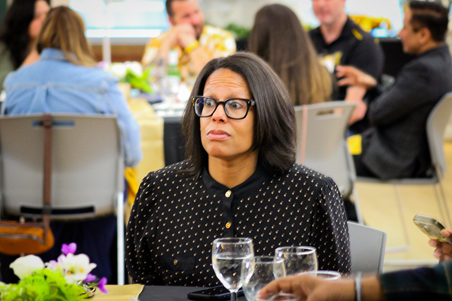 HILP Dinner 2025-2026 / A woman with glasses and a patterned blouse sits at a table in a busy room, looking thoughtful. There are flowers and water glasses on the table.