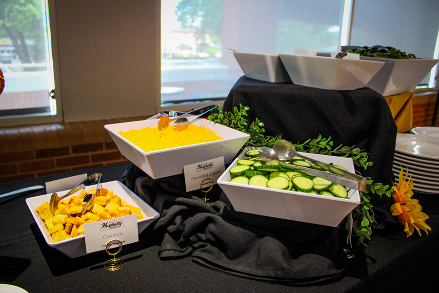HILP Dinner 2025-2026 / Buffet table with white bowls containing croutons, shredded cheese, and sliced cucumbers. Displayed on black cloth with greenery for decoration.