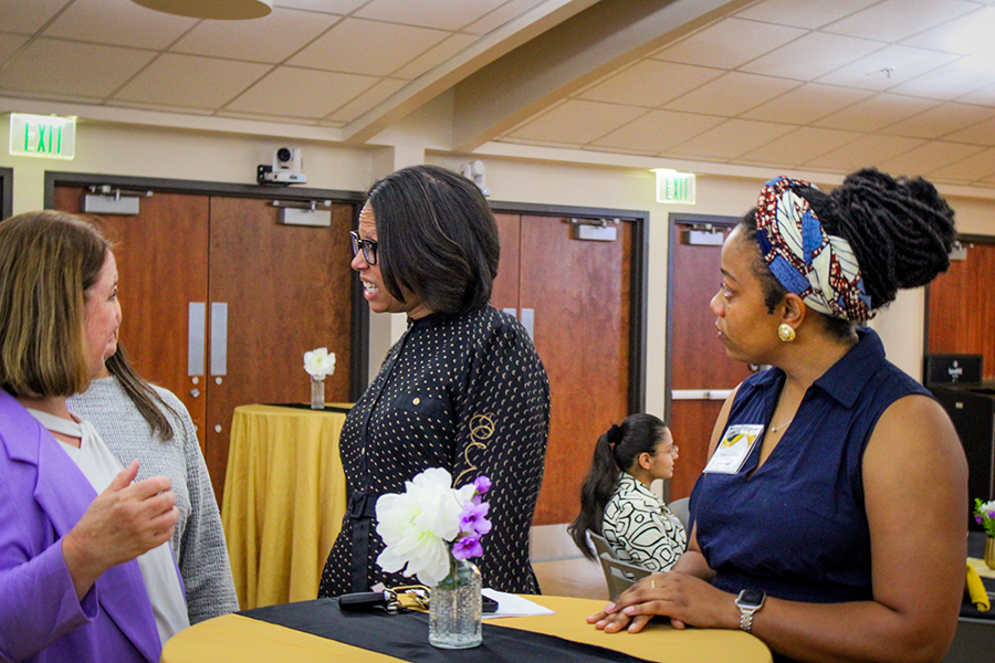 HILP Dinner 2025-2026 / Three women engage in conversation at a social event, with a table holding a vase of flowers nearby. The mood is friendly and interactive.
