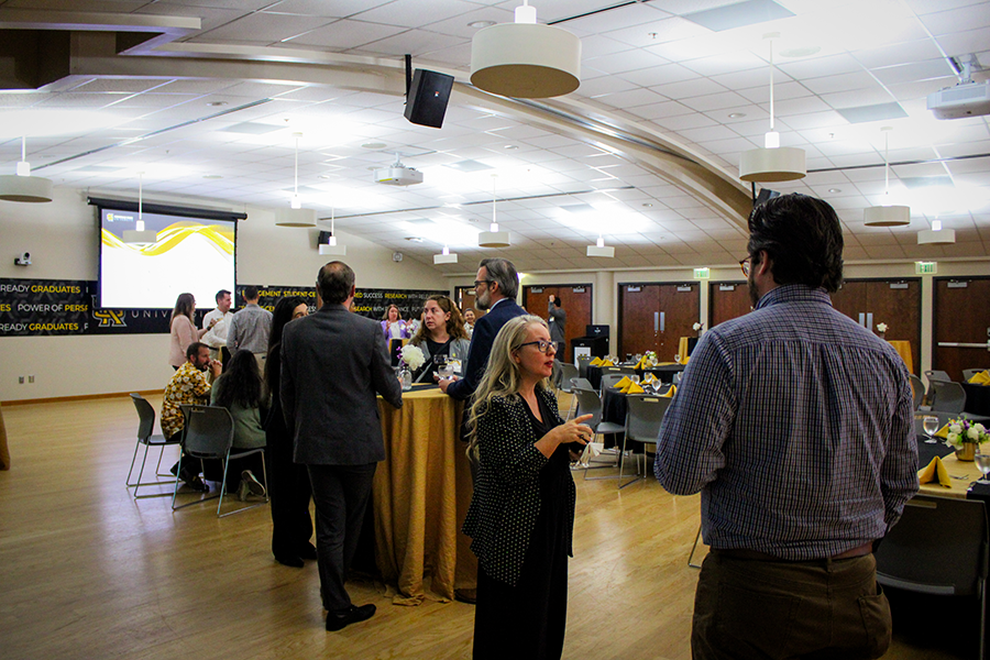 HILP Dinner 2025-2026 / A group of people socialize in an event hall with round tables covered in yellow tablecloths. A projector displays a presentation on the wall.