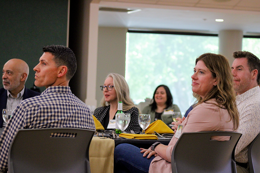 HILP Dinner 2025-2026 / A group of attentive people sit around a table at a casual indoor event. Bright daylight filters through large windows, creating a warm, engaged atmosphere.