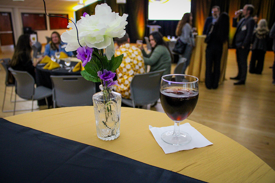 HILP Dinner 2025-2026 / A glass of red wine on a cocktail table with a white and purple flower arrangement in a clear vase. People socialize in a warmly lit banquet setting.