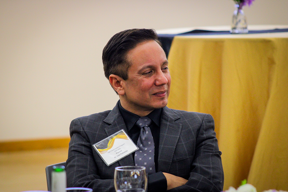 HILP Dinner 2025-2026 / Man in a dark suit with a name tag smiles in profile at an event, seated at a table with a glass in the foreground and a yellow tablecloth behind.