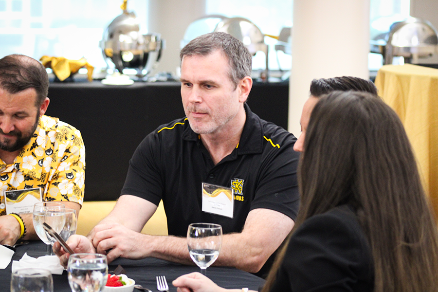 HILP Dinner 2025-2026 / Middle-aged man in a black and yellow shirt sits attentively at a round table during a formal event. Around him are others, dressed for the occasion, included in thoughtful conversation.