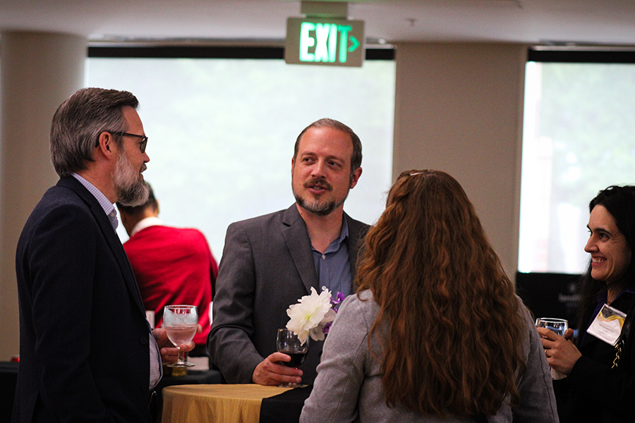 HILP Dinner 2025-2026 / Four people engage in a lively conversation at a networking event, holding drinks. They're gathered around a table with flowers, under a lit "Exit" sign.