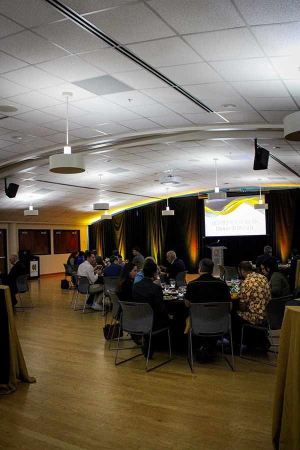 HILP Dinner 2025-2026 / A well-lit banquet hall with people seated at round tables, engaging in conversation. A large screen displays an event title. The ambiance is formal and inviting.