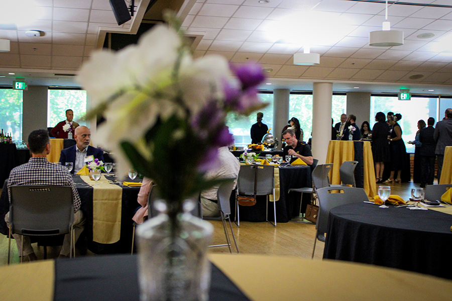 HILP Dinner 2025-2026 / A clear vase with white and purple flowers sits on a table at a social event. Blurred background shows people gathered around tables.