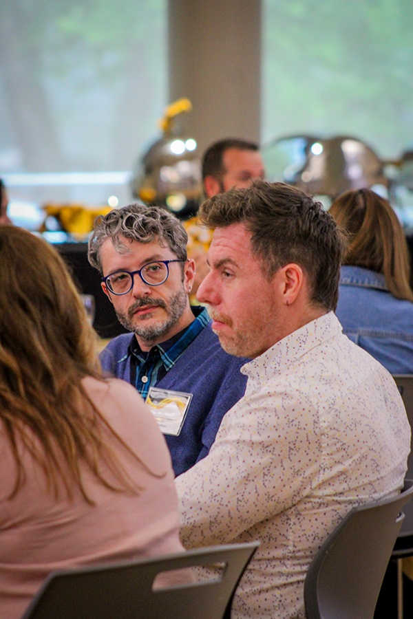HILP Dinner 2025-2026 / Two men in thoughtful conversation at an indoor event, seated among others. The setting is casual with blurred backgrounds of tables and windows.