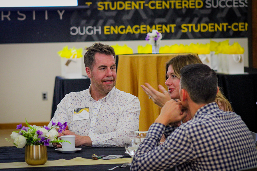 HILP Dinner 2025-2026 / Three people engaged in conversation at a table decorated with flowers. The setting appears to be an event focused on student engagement and success.