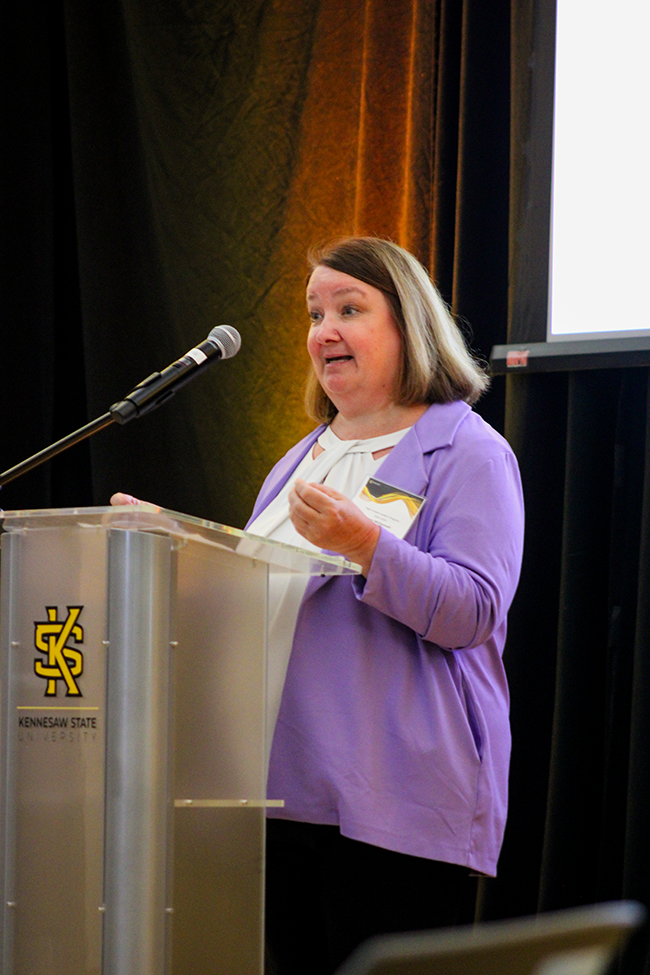 HILP Dinner 2025-2026 / A woman in a purple blazer speaks at a podium with a microphone, bearing the Kennesaw State University logo. She appears engaged and focused.