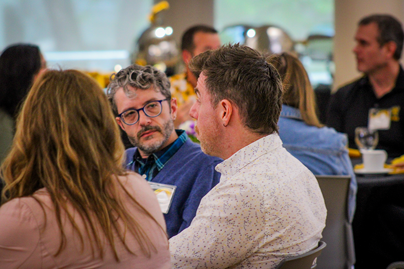 HILP Dinner 2025-2026 / A group of people seated at a table engaged in conversation at a social event. The setting is relaxed with a backdrop of blurred decorations. The mood appears lively and engaged.