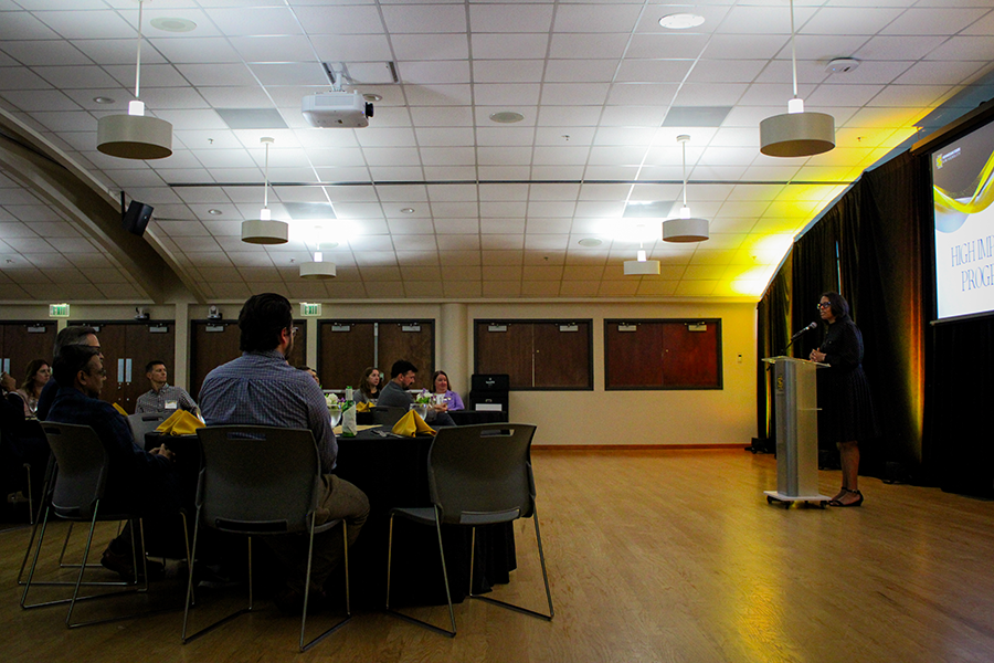 HILP Dinner 2025-2026 / A group of people sit at round tables with black tablecloths and yellow napkins, attentively listening to a speaker on stage in a warmly lit banquet hall.