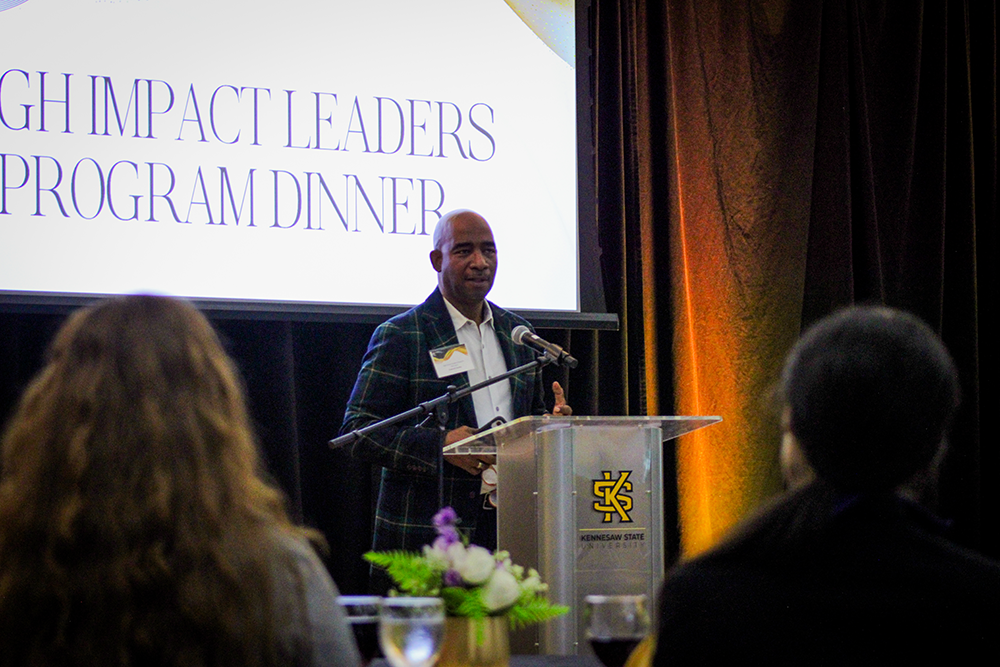 HILP Dinner 2025-2026 / A speaker stands at a podium with "Impact Leaders Program Dinner" displayed on a screen. The setting is formal, with attendees seated at tables.