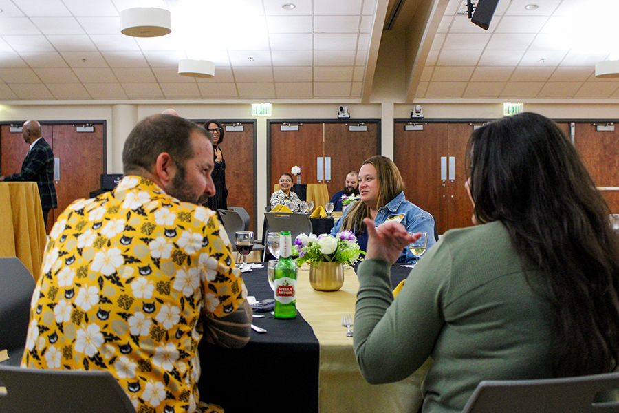 HILP Dinner 2025-2026 / A group of people sit at a banquet table covered with a yellow tablecloth, chatting and smiling. Green bottles and a floral centerpiece add to the festive atmosphere.