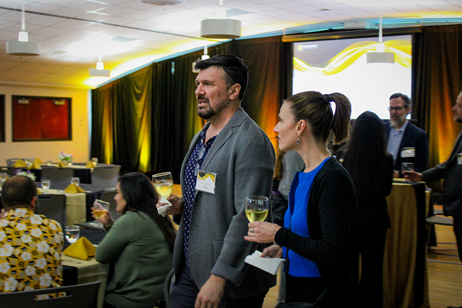 HILP Dinner 2025-2026 / A man and woman holding wine glasses converse at an event with elegantly set tables and warm lighting, creating a sophisticated and lively atmosphere.