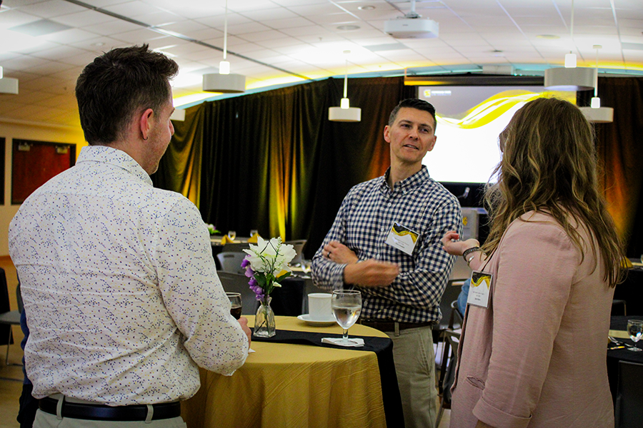 HILP Dinner 2025-2026 / Three people engage in conversation at a formal event. They stand around a table with a gold cloth. The room is warmly lit, with a backdrop of black curtains.