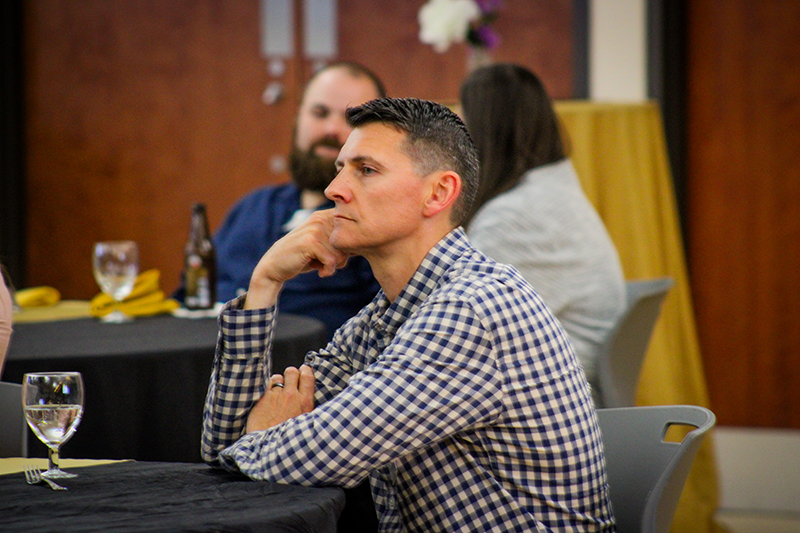 HILP Dinner 2025-2026 / A man in a checkered shirt sits thoughtfully at a table with folded arms, surrounded by other seated individuals at an event, conveying a reflective mood.