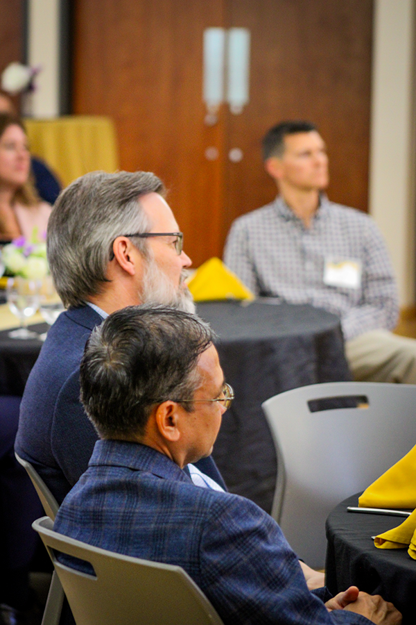 HILP Dinner 2025-2026 / Men in suits attentively listen during a formal event. They are seated at tables with yellow napkins, conveying a professional atmosphere.