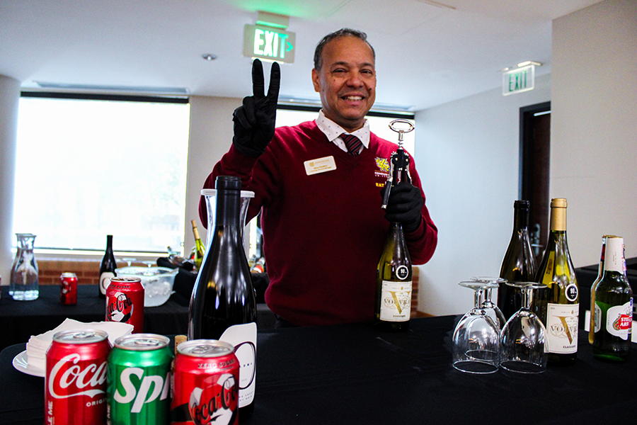 HILP Dinner 2025-2026 / A smiling bartender in a red sweater flashes a peace sign while holding a wine bottle and corkscrew. Assorted drinks and glasses are on the table.