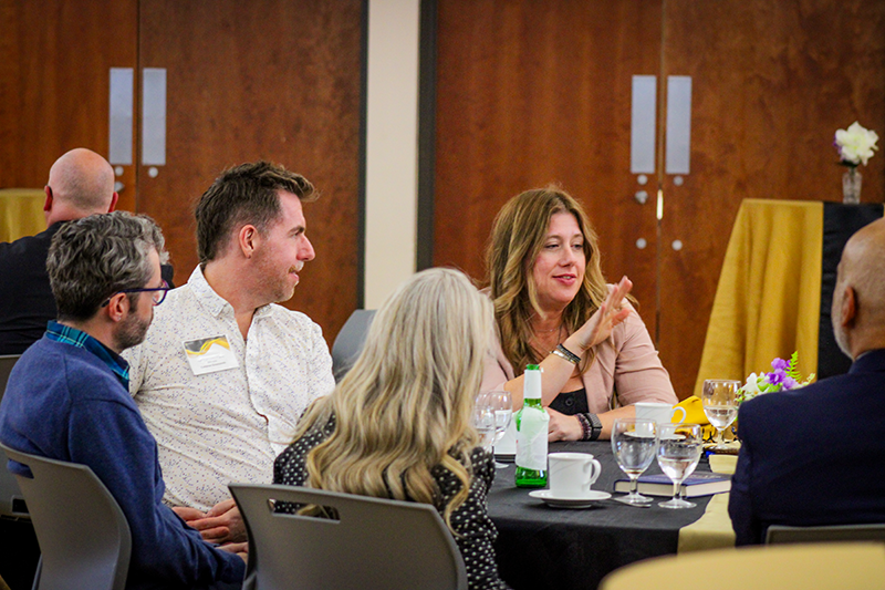 HILP Dinner 2025-2026 / A group of people sit around a table at an event, engaged in conversation. The setting is formal, with glasses, drinks, and yellow table decor. The mood is lively.