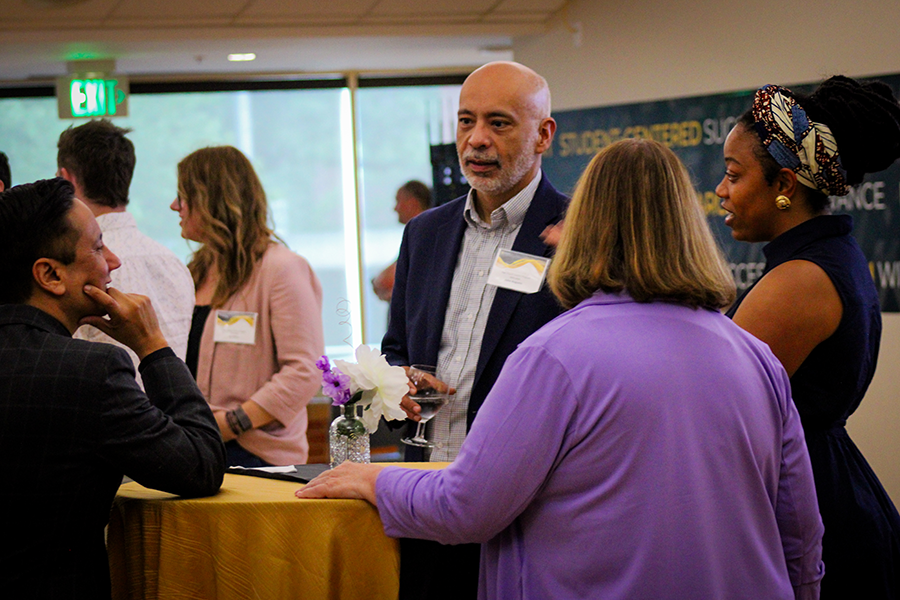 HILP Dinner 2025-2026 / A group of people engage in conversation at a networking event. They stand around a table with flowers, looking engaged and relaxed.