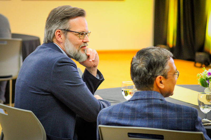HILP Dinner 2025-2026 / Two men in suits sit at a round table with black tablecloths, engaged in conversation. A vase of flowers and glasses of water are in the foreground.