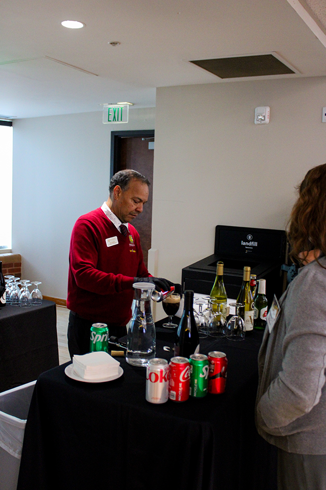 HILP Dinner 2025-2026 / A man in a red sweater pours a drink at a beverage station with wine bottles, soda cans, and glassware. Another person waits. The setting is casual and organized.