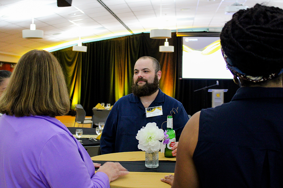 HILP Dinner 2025-2026 / A man engaged in conversation stands at a table with a woman in purple and another with braided hair. The venue is dimly lit with black and gold accents, giving an elegant, formal atmosphere.