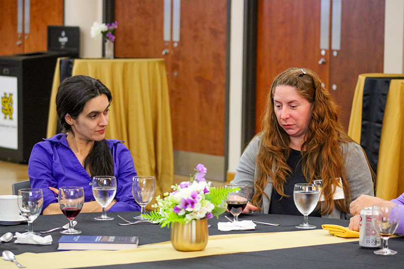 HILP Dinner 2025-2026 / Two women seated at a banquet table with folded arms, appearing thoughtful. The table has wine glasses and a floral centerpiece, set in a formal room.