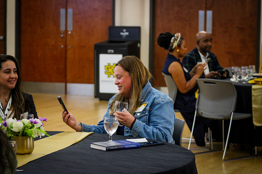 HILP Dinner 2025-2026 / A woman in a denim jacket smiles at her phone while holding a glass, seated at a table with flowers and books. Others chat at a nearby table. Casual, friendly atmosphere.