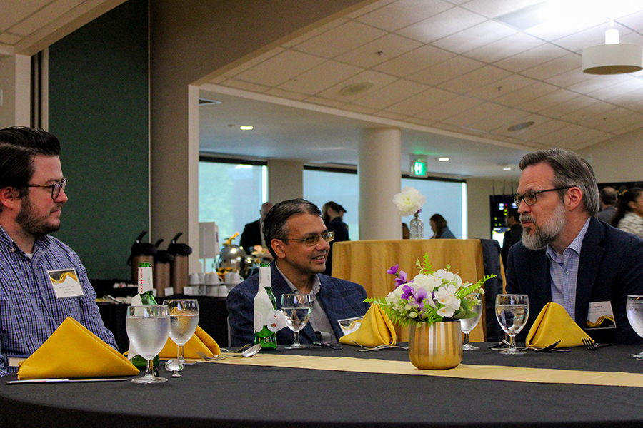 HILP Dinner 2025-2026 / Three men in business attire sit around a table with yellow napkins and a flower centerpiece, engaged in conversation in a formal setting.