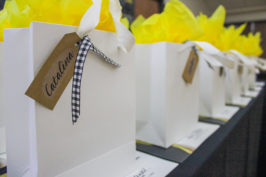 HILP Dinner 2025-2026 / Rows of white gift bags with bright yellow tissue paper and name tags, neatly arranged on a black table in a softly lit room, suggest a celebratory or formal event.