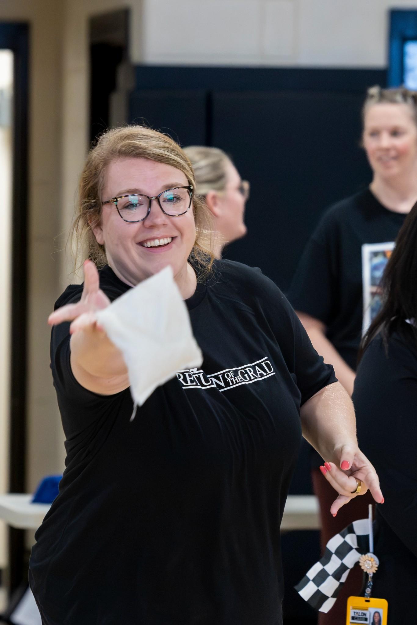 2026 Owlympics / Smiling woman in glasses gestures while tossing a bean bag at Owlympics, surrounded by people in a lively indoor gathering; playful, welcoming mood.