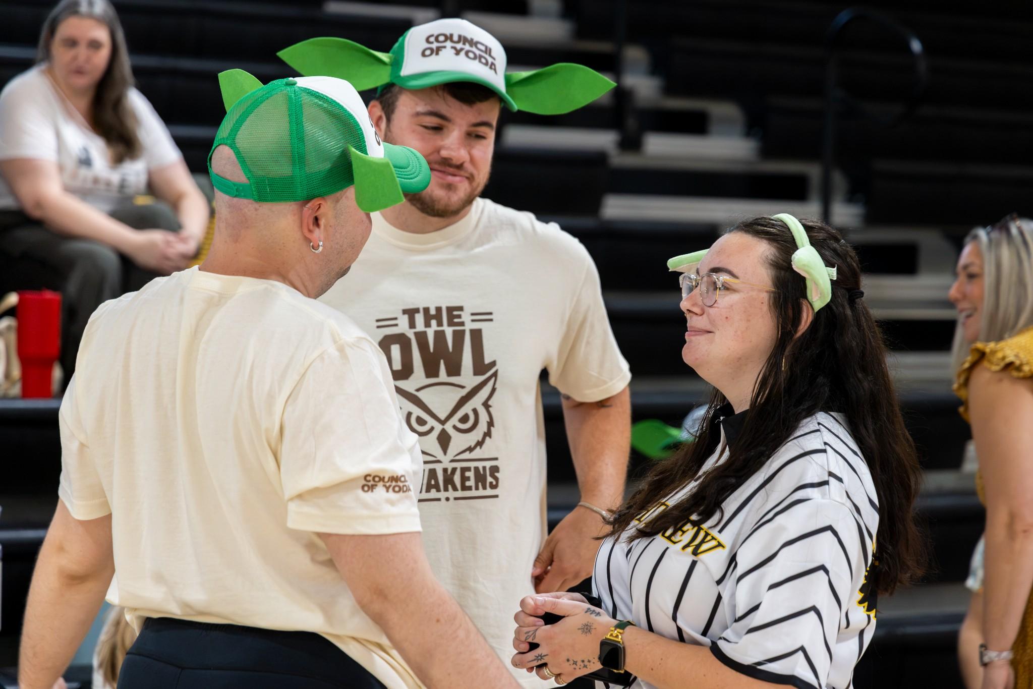 2026 Owlympics / Three smiling fans in owl-themed shirts and grogu caps chat courtside at a lively sports event, creating a cheerful atmosphere.