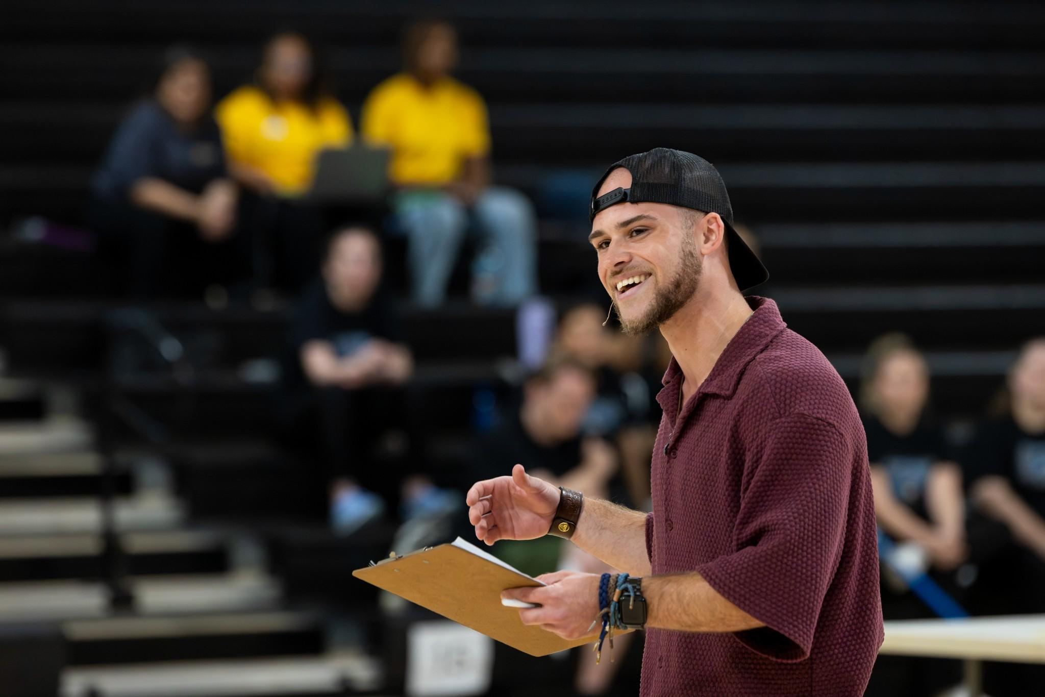 2026 Owlympics / Smiling young man in a backward cap and maroon shirt holds a clipboard before a blurred audience, creating an upbeat, engaging atmosphere.