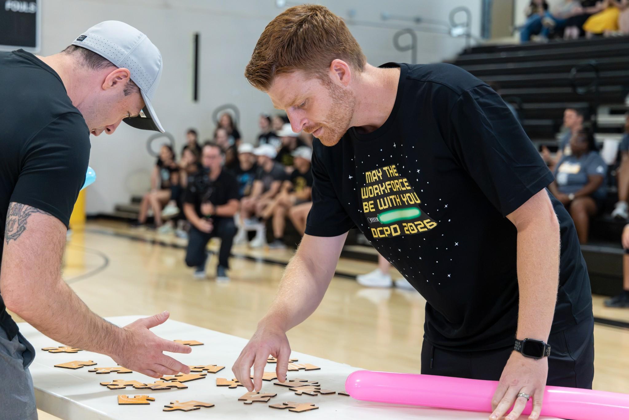 2026 Owlympics / Two men compete in a gym puzzle challenge, arranging wooden pieces on a table as spectators watch; focused, energetic atmosphere.