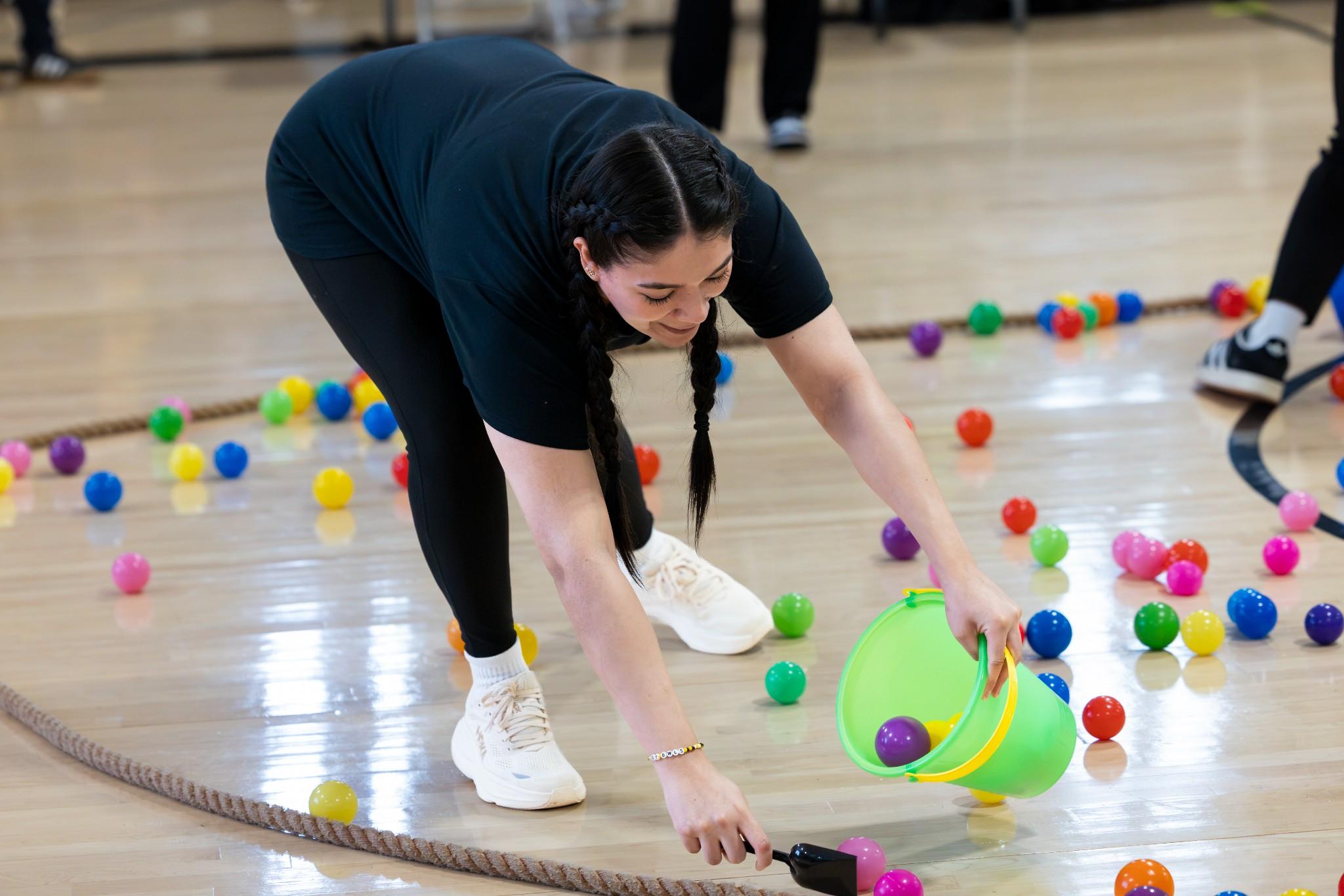 2026 Owlympics / Woman in black athletic wear gathers colorful balls with a green bucket during an Owlympics game, surrounded by ropes and scattered balls.