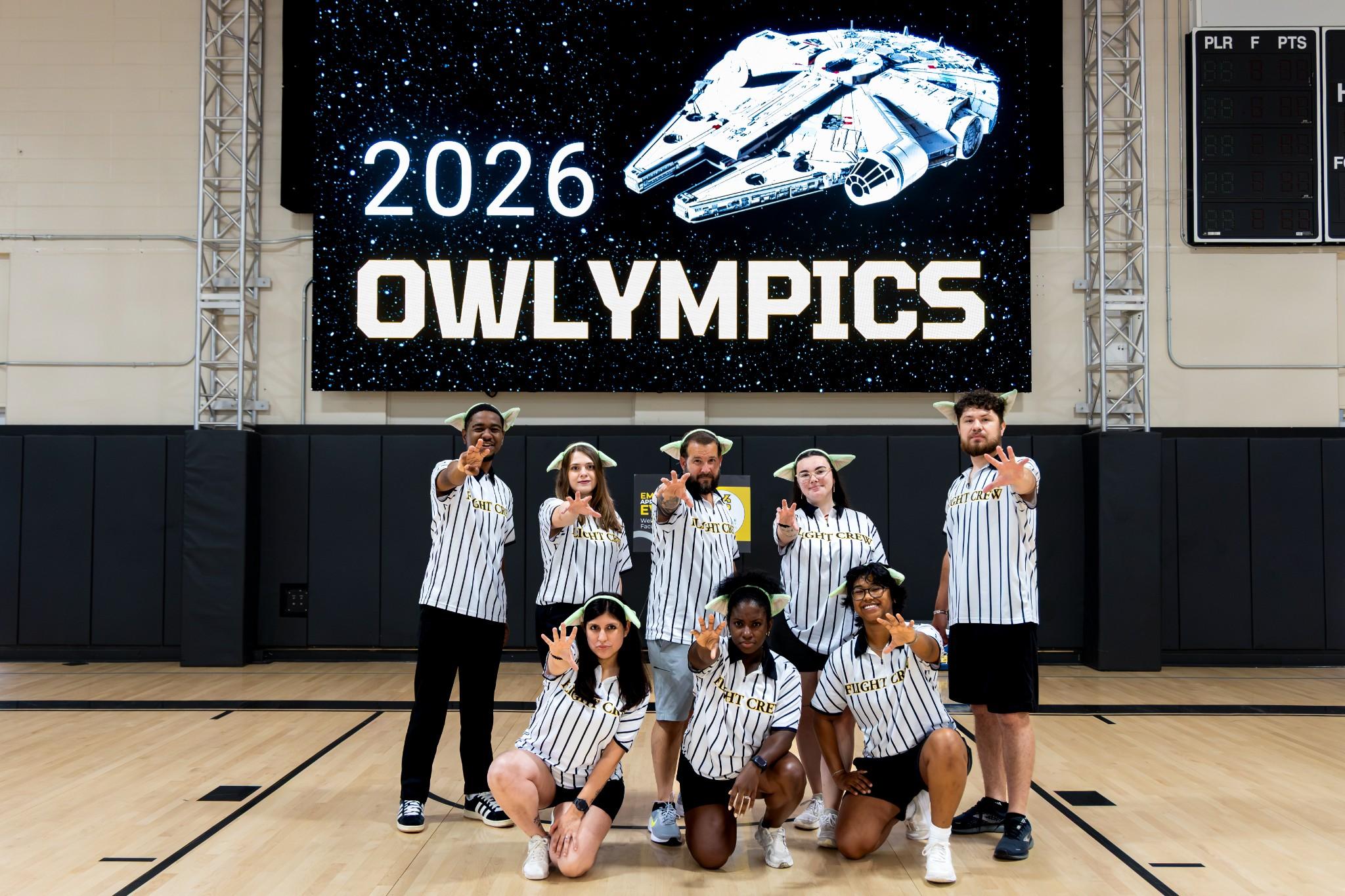 2026 Owlympics / Eight-person group poses on a basketball court beneath a “2026 OWLympics” banner with a Star Wars spaceship, conveying team spirit and excitement.