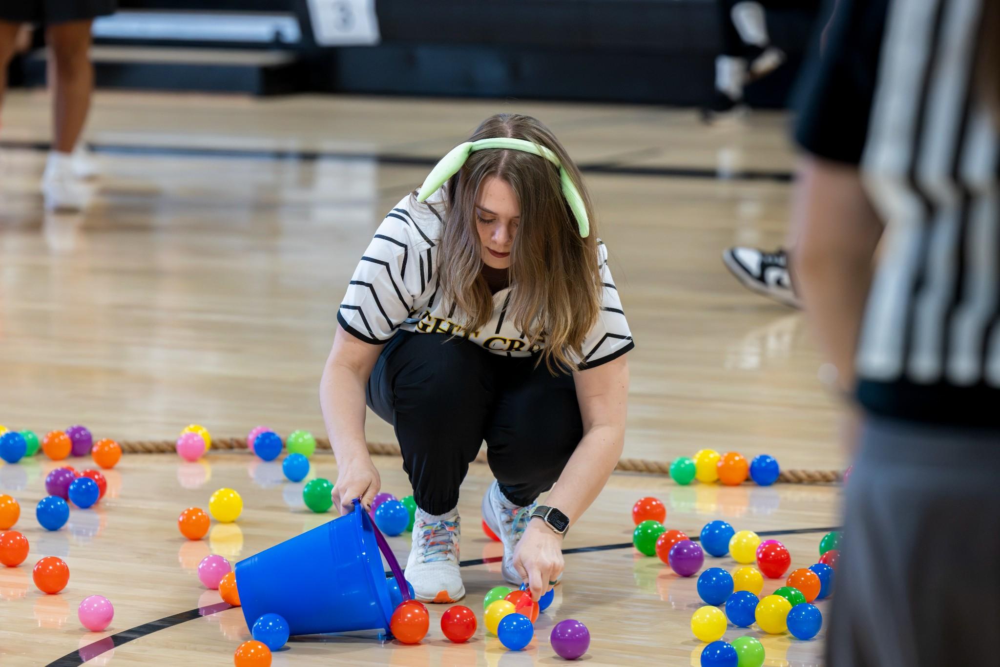 2026 Owlympics / Young woman in a gym gathers colorful plastic balls into a blue bucket, while referees stand nearby; playful, energetic scene.