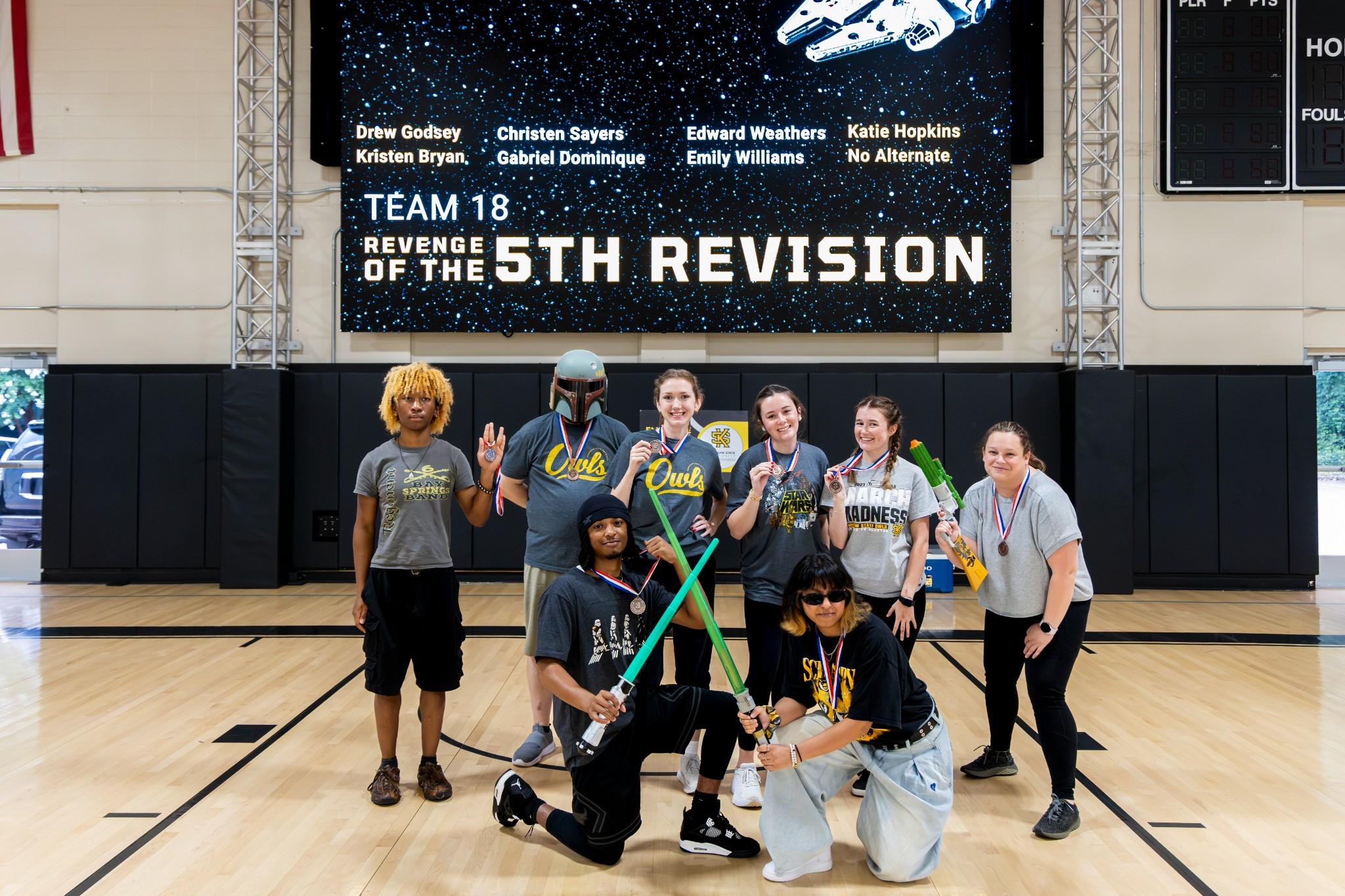 2026 Owlympics / Eight smiling people pose on a basketball court beneath a “Team 18: Revenge of the 5th Revision” banner, celebrating teamwork.