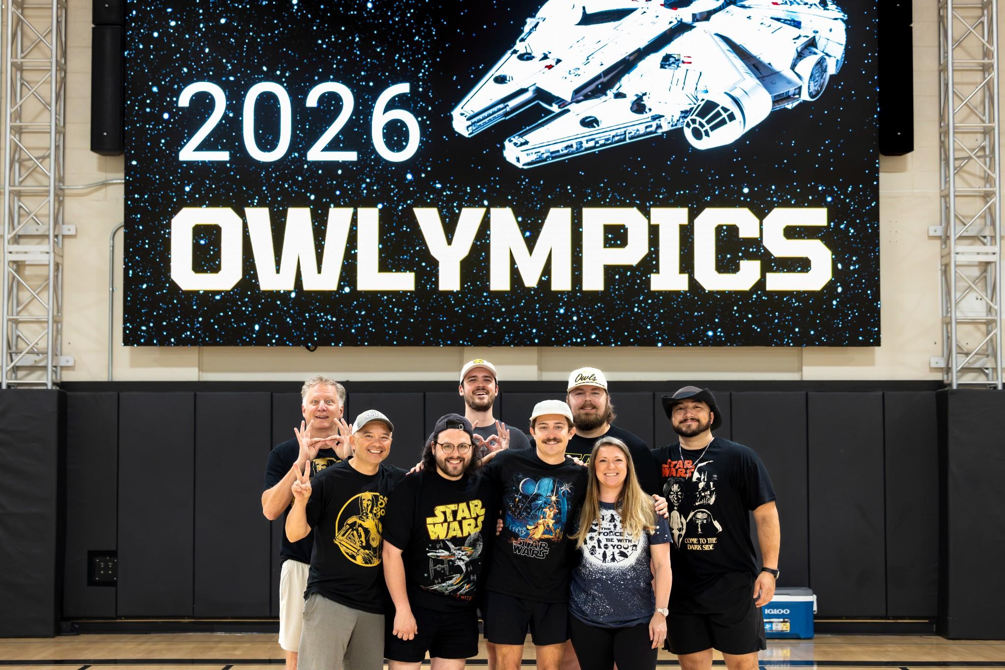 2026 Owlympics / A smiling group poses beneath a “2026 OWLYMPICS” Star Wars banner featuring a spaceship, creating a festive, sci-fi convention atmosphere.
