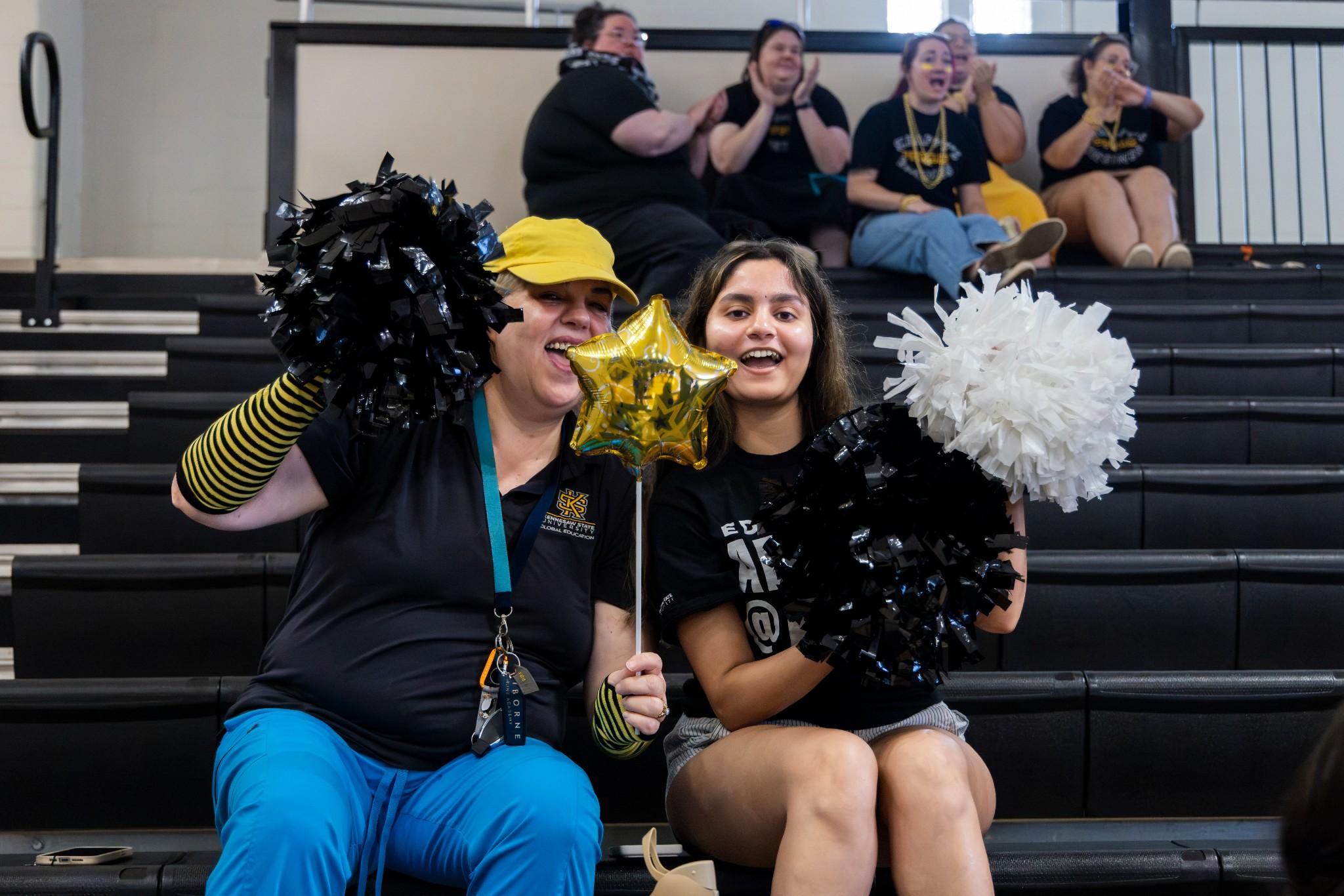 2026 Owlympics / Two joyful fans pose in bleachers with black and white pom-poms and a gold star wand, surrounded by cheering supporters.