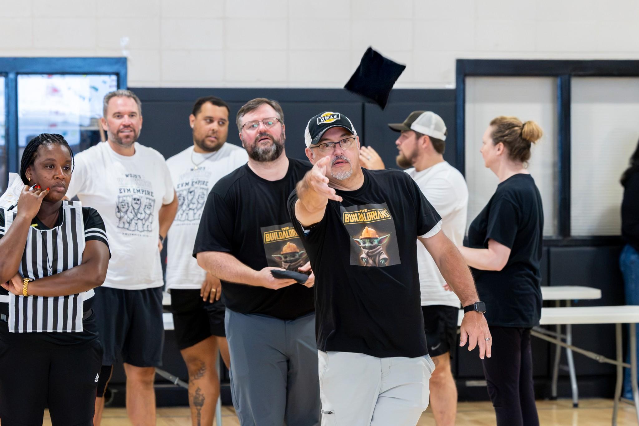 2026 Owlympics / Team in a gym watches a bearded man in a cap toss a black beanbag, while a referee and others look on with amused curiosity.
