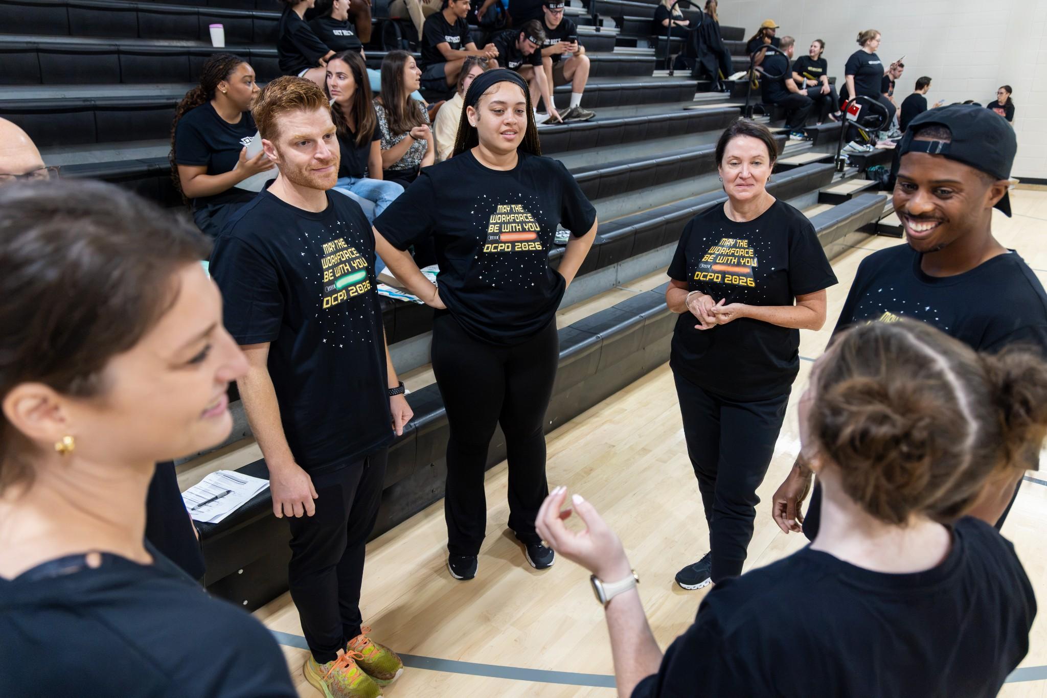 2026 Owlympics / Diverse adults in matching black shirts gather on a gym court, smiling and talking near bleachers in a warm, welcoming team setting.