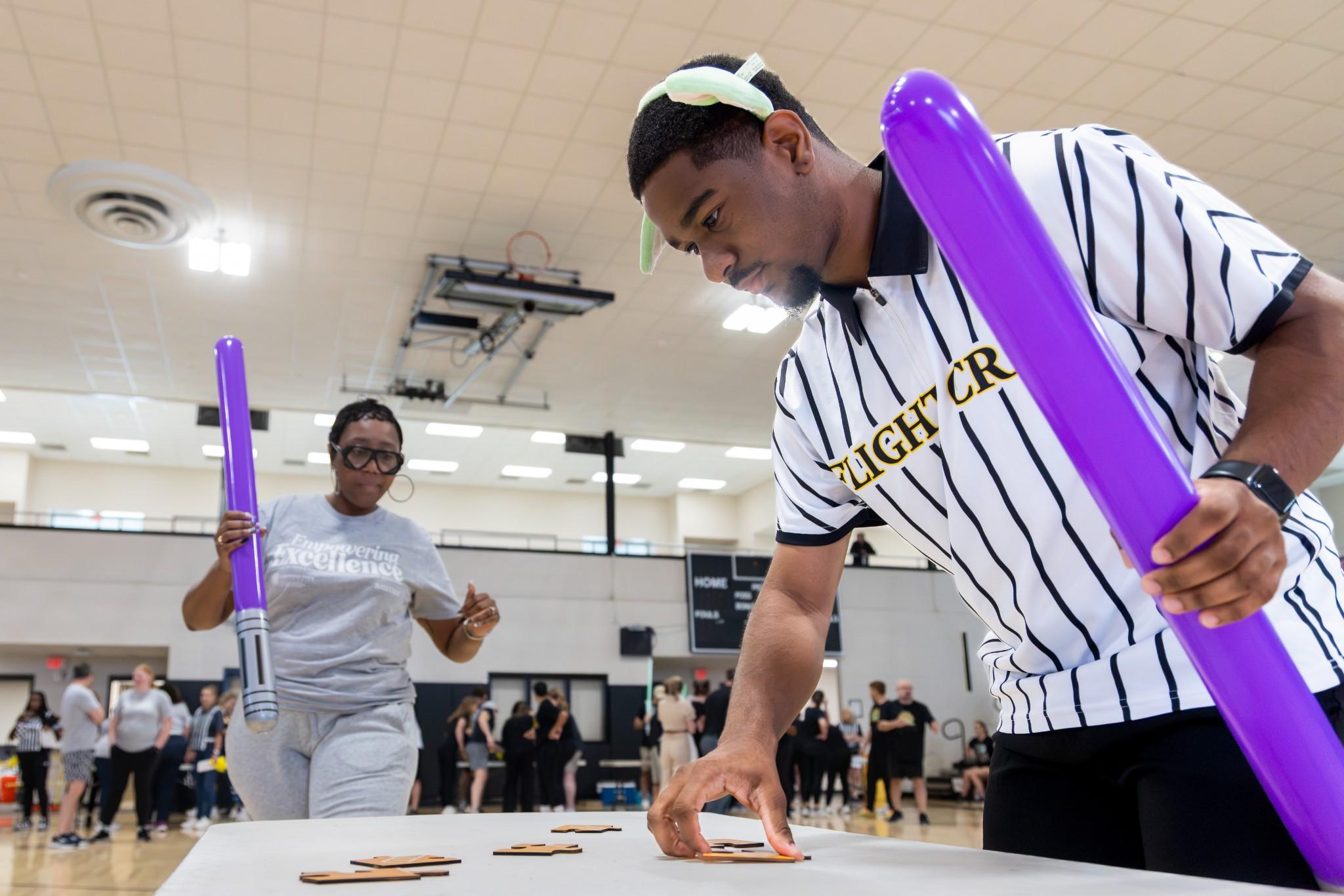 2026 Owlympics / Two adults play an energetic indoor table game with purple light sabers, moving puzzle pieces across a table as spectators watch in a gym.