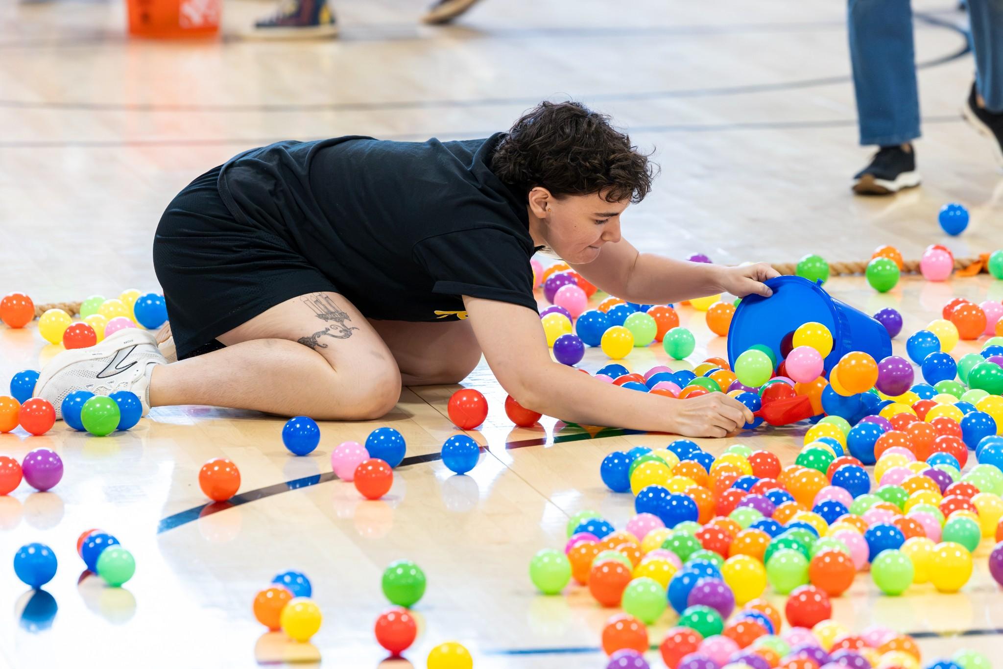 2026 Owlympics / Person in black clothing crawls across a gym floor, reaching through a colorful sea of plastic balls during a playful, energetic activity.