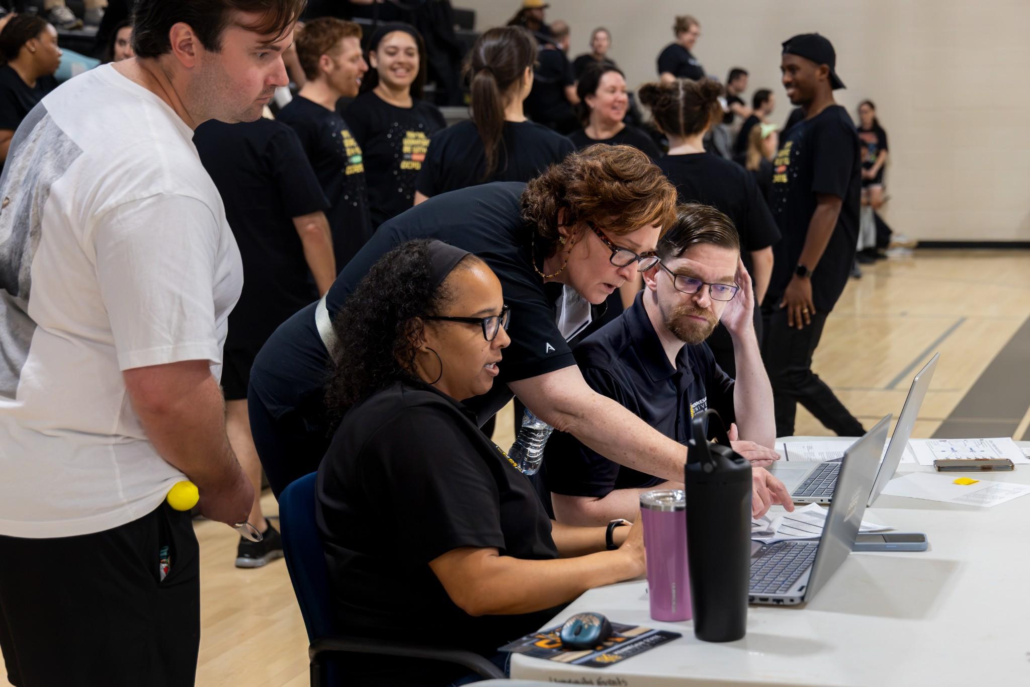 2026 Owlympics / At a busy indoor event, three volunteers collaborate at a laptop and paperwork while a crowd gathers behind them in a lively gym.